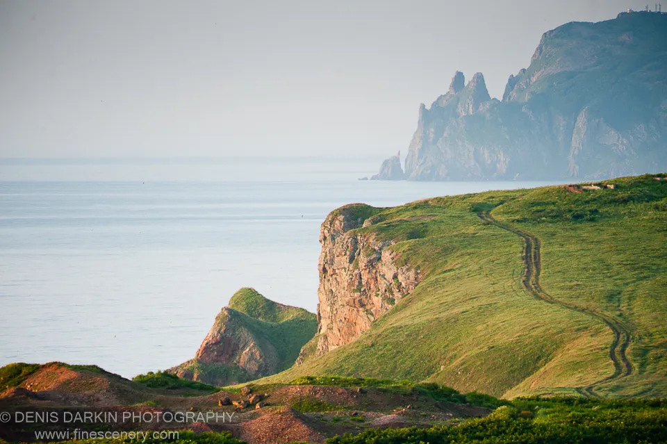 A winding dirt track cuts through green coastal cliffs at Topauza Bay, dropping toward the calm sea with jagged rocky peaks rising in the distance under pale sky.