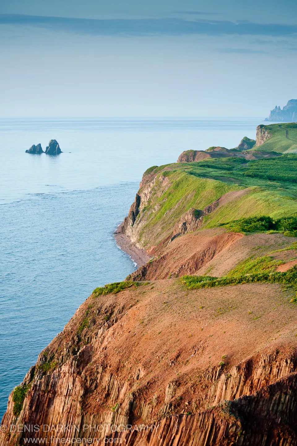 Columnar rock cliffs with terraced green slopes descend to calm water at Topauza Bay, a solitary sea stack marking the far horizon under hazy sky.