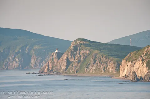 A white lighthouse stands on a steep headland at Topauza Bay, surrounded by muted green and tan coastal cliffs curving gently into the calm sea below.