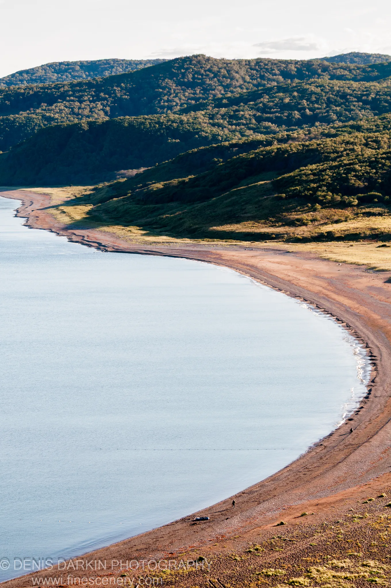 Lush green hills rise steeply behind a sandy pebbly beach, long shadows cast by low-angle light on the Samarga River bank, early autumn.