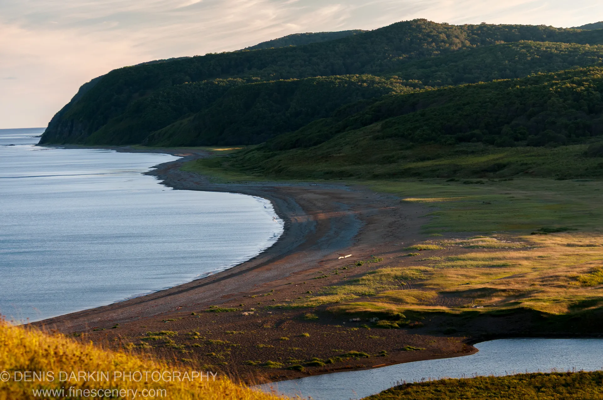Calm water meets a pebbly beach below dark forested hills, at the Samarga River mouth in early autumn, golden-hour sky above.