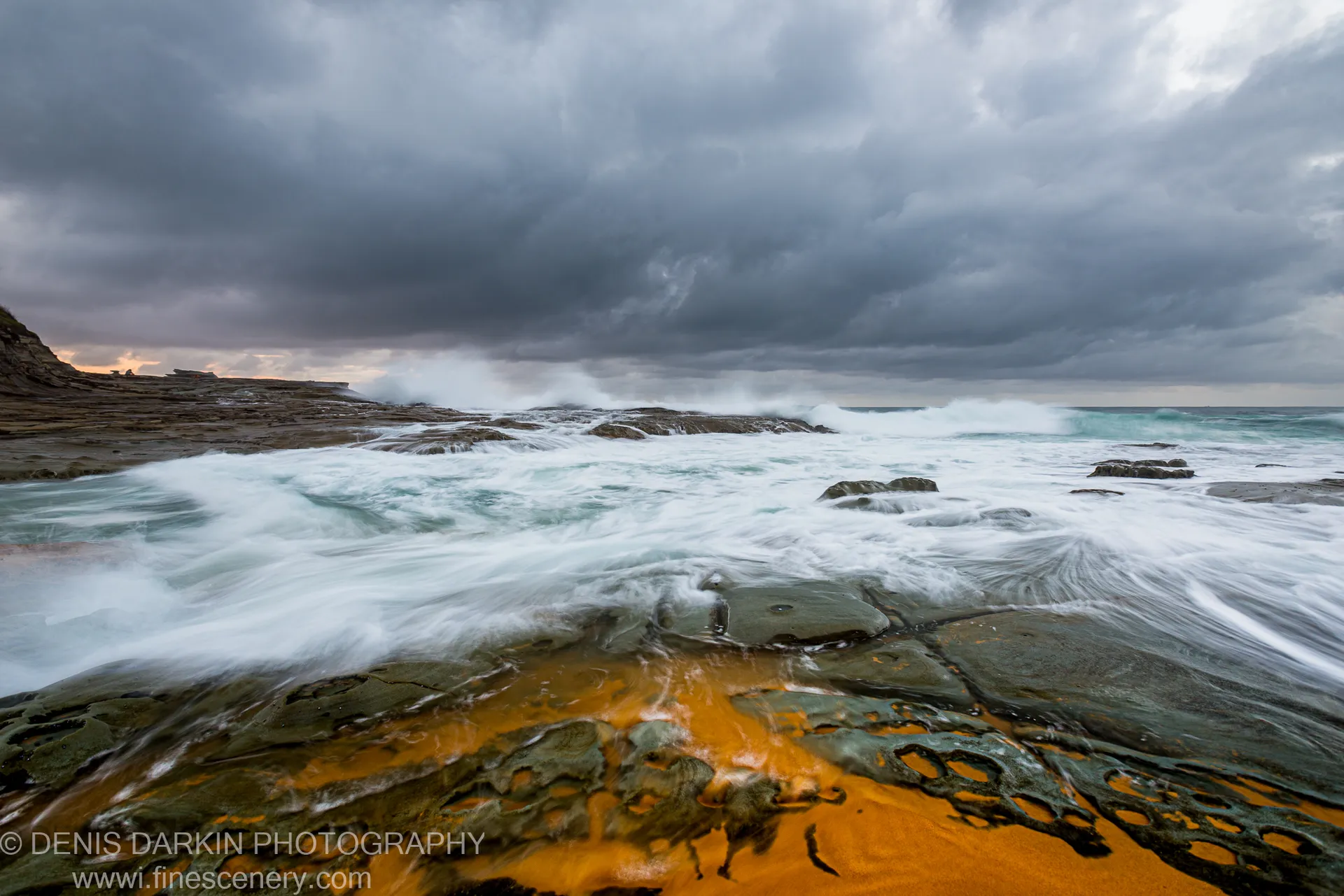 Iron-stained sandstone pools reflect orange light on Spoon Bay's rock platform at dawn, with storm clouds overhead.