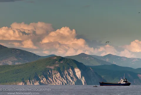 A blue cargo ship at anchor near a small boat, with limestone cliffs, forest, and cumulus clouds at golden hour, Dva Brata Islands, Primorsky Krai.
