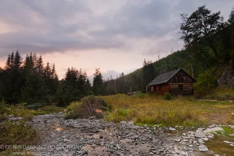 A rustic log cabin sits beside a rocky stream at the foot of a forested slope in St Helen Springs, Primorsky Krai.