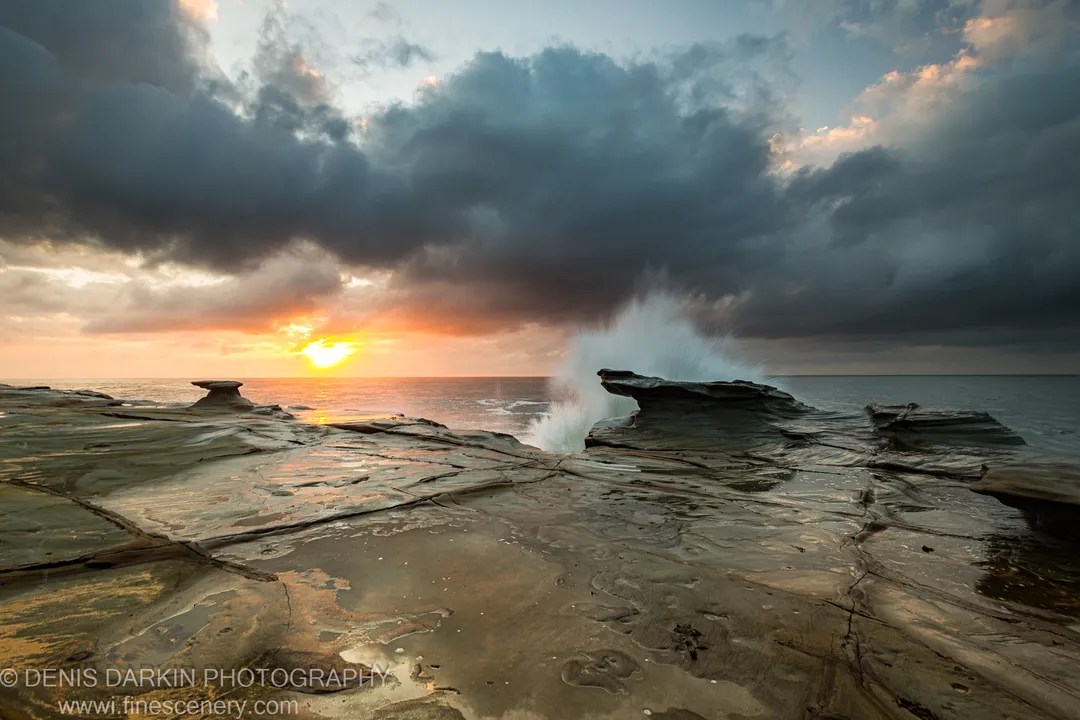 A wave breaks against a sandstone ledge at Spoon Bay, freezing its white spray against storm clouds at dawn.