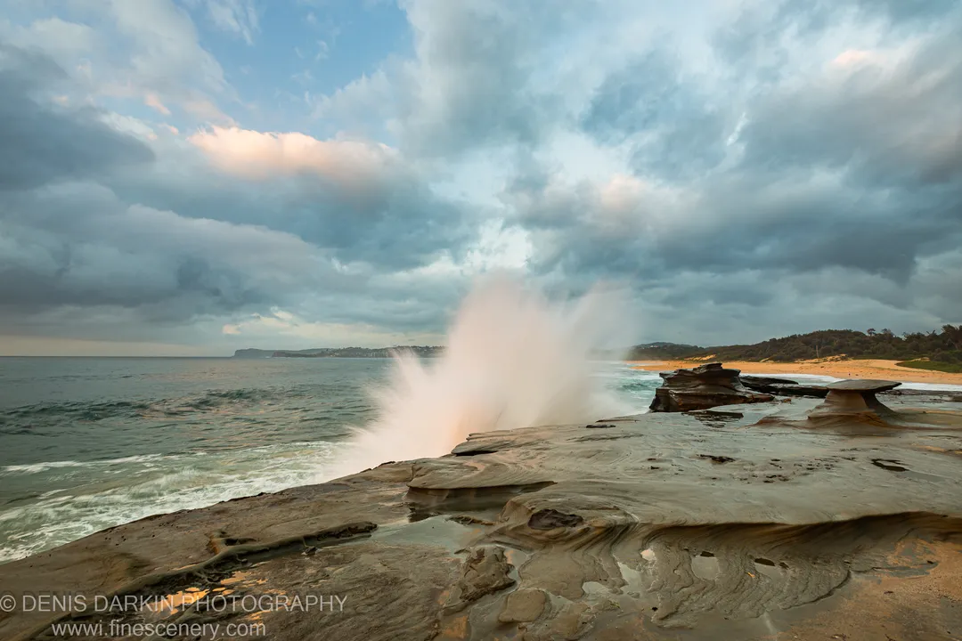 A wave erupts into a tall plume of spray against dark storm clouds above the Spoon Bay sandstone platform at dawn.