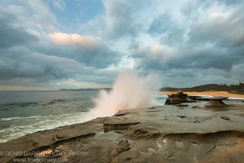A wave erupts into a tall plume of spray against dark storm clouds above the Spoon Bay sandstone platform at dawn.