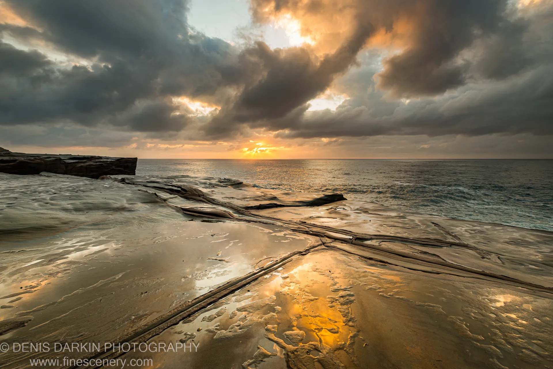 Rock pools on a sandstone shelf at Spoon Bay reflect the bright horizon light beneath dark storm clouds at dawn.