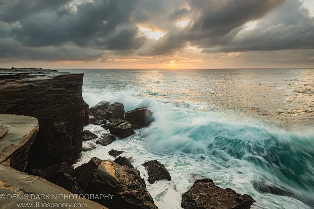 Long-exposure waves break across a flat rock platform at Spoon Bay under heavy storm clouds at dawn.