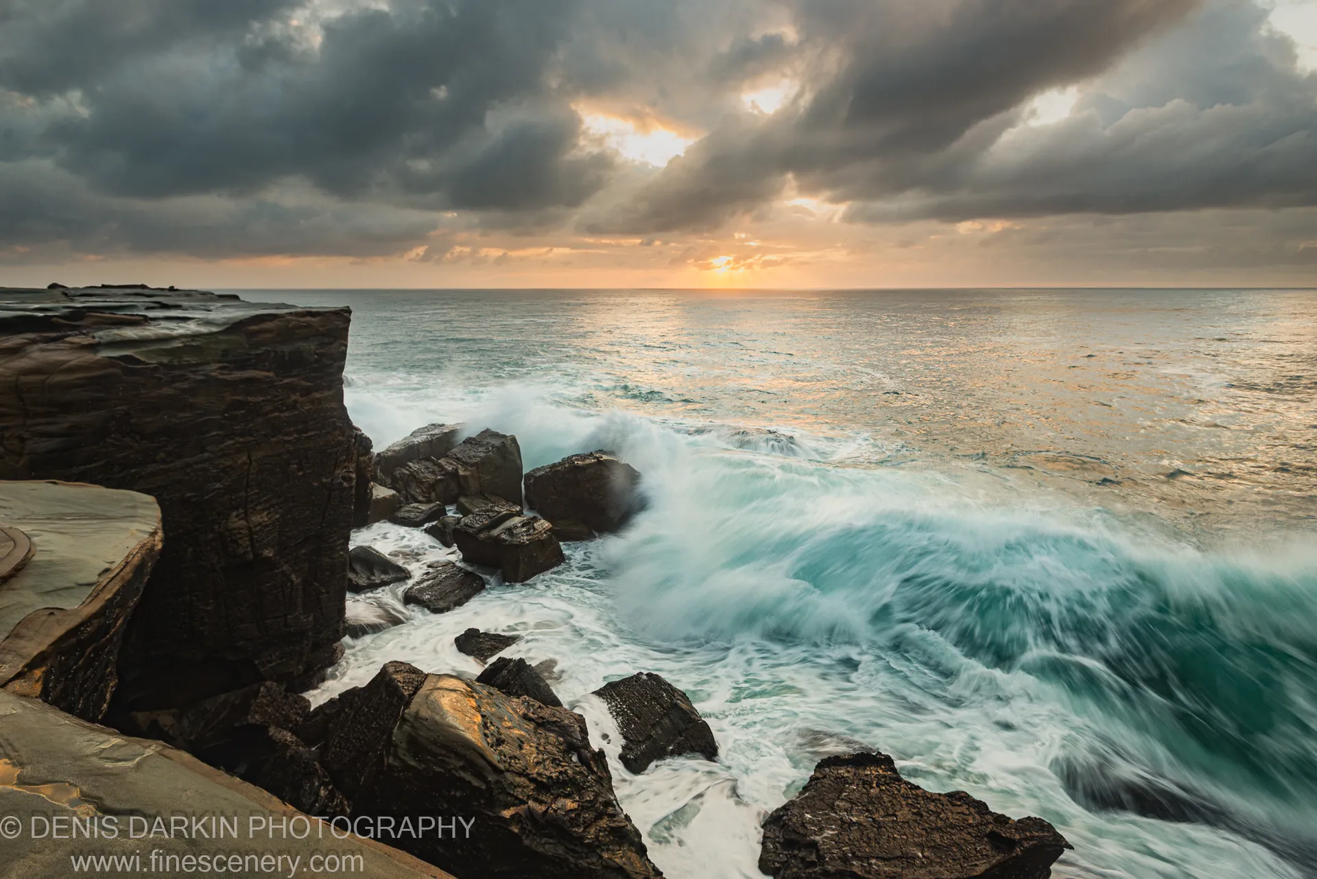 Long-exposure waves break across a flat rock platform at Spoon Bay under heavy storm clouds at dawn.