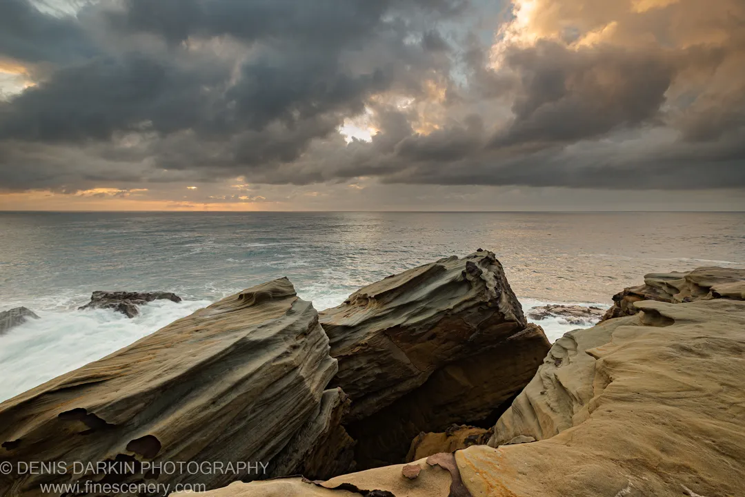 Layered sandstone shelves at Spoon Bay at dawn, with heavy storm clouds overhead and warm sunlight on the ocean.