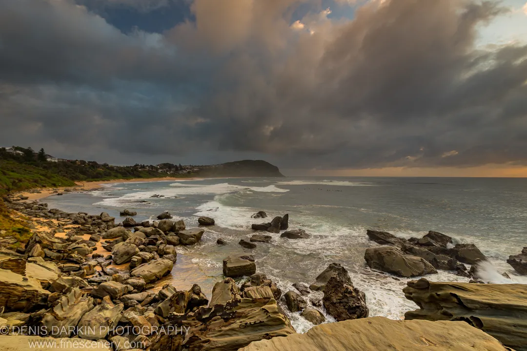 A sandy beach curves along Spoon Bay toward a dark headland under storm clouds at dawn, New South Wales.