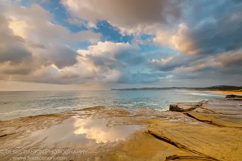Wet sandstone rock planes at Spoon Bay mirror the cloud formations above at dawn, with warm light on the stone edges.