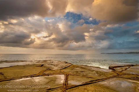Layered sandstone with deep fissures at Spoon Bay, lit by low-angle dawn light under a sky of clouds and ocean beyond.