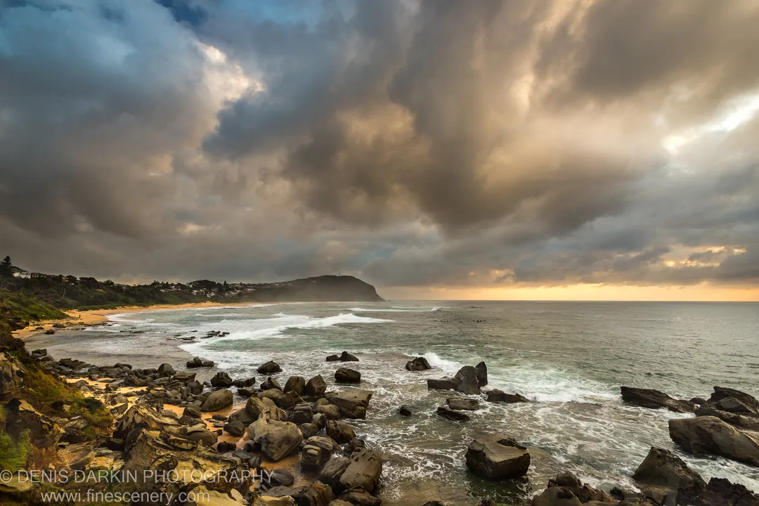 Heavy grey clouds over Spoon Bay lit by warm golden light from the right horizon on a summer morning.