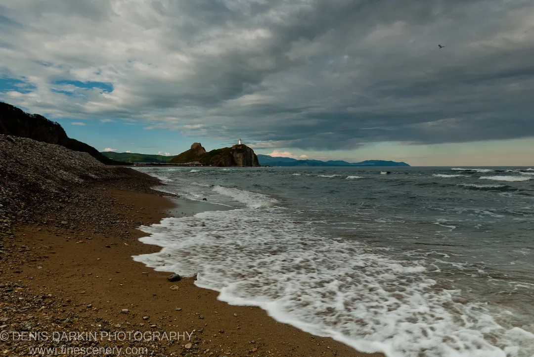 White lighthouse on a rocky headland under breaking storm clouds, with a sandy beach and waves in the foreground at Dva Brata Islands, Russia.