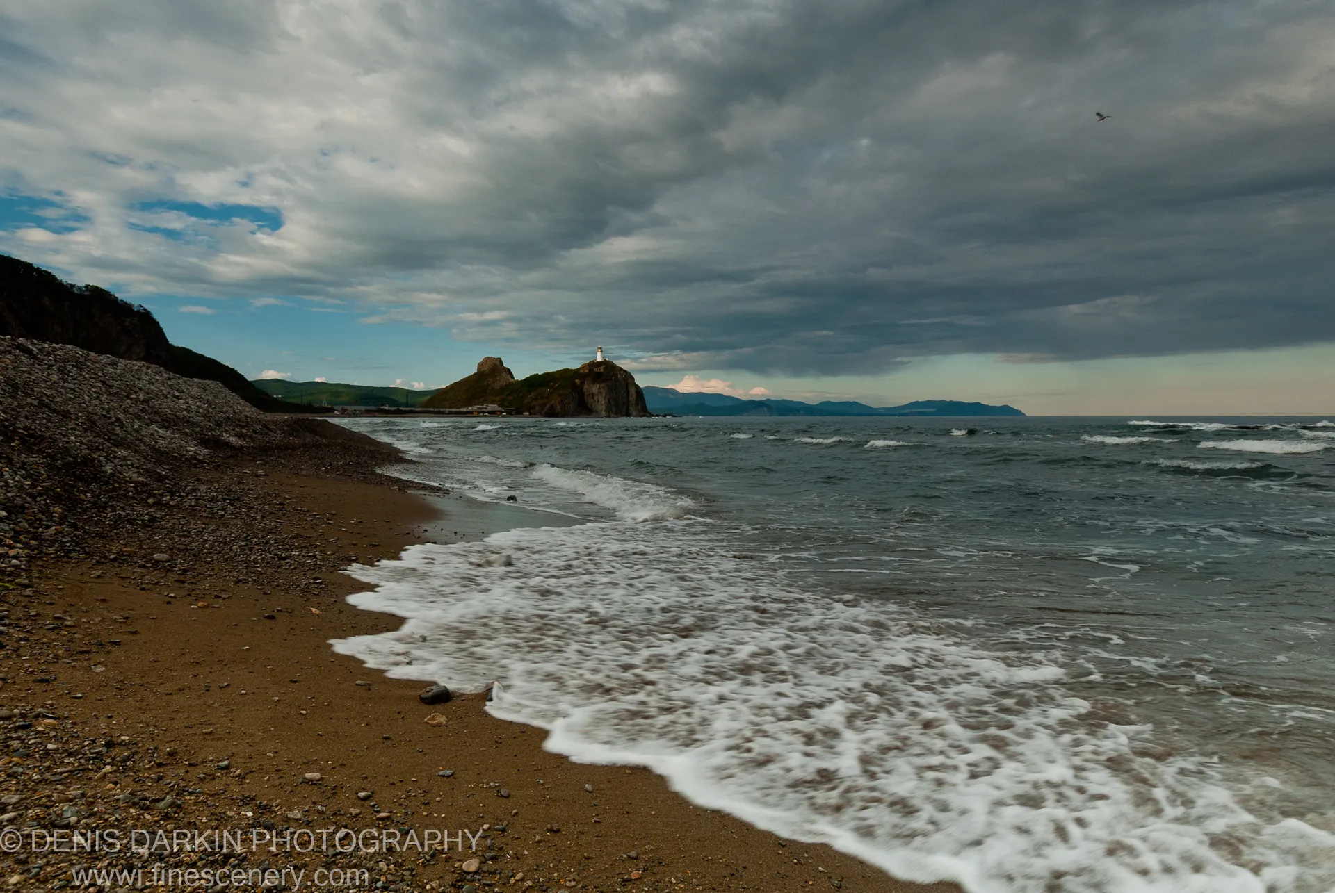 White lighthouse on a rocky headland under breaking storm clouds, with a sandy beach and waves in the foreground at Dva Brata Islands, Russia.