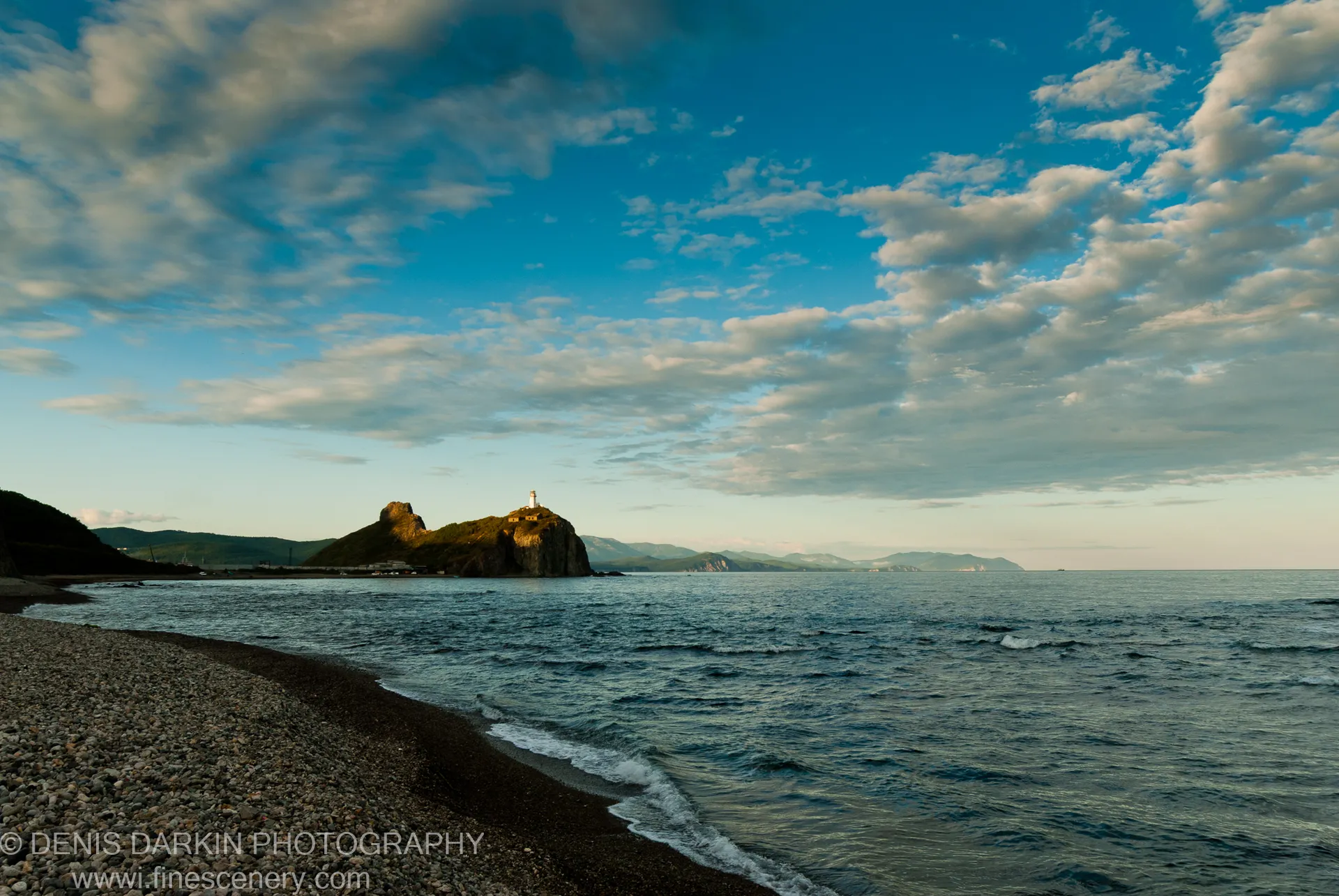 Dark pebble shoreline curving toward a grass-covered headland with a white lighthouse, Terneysky coast, scattered clouds above.