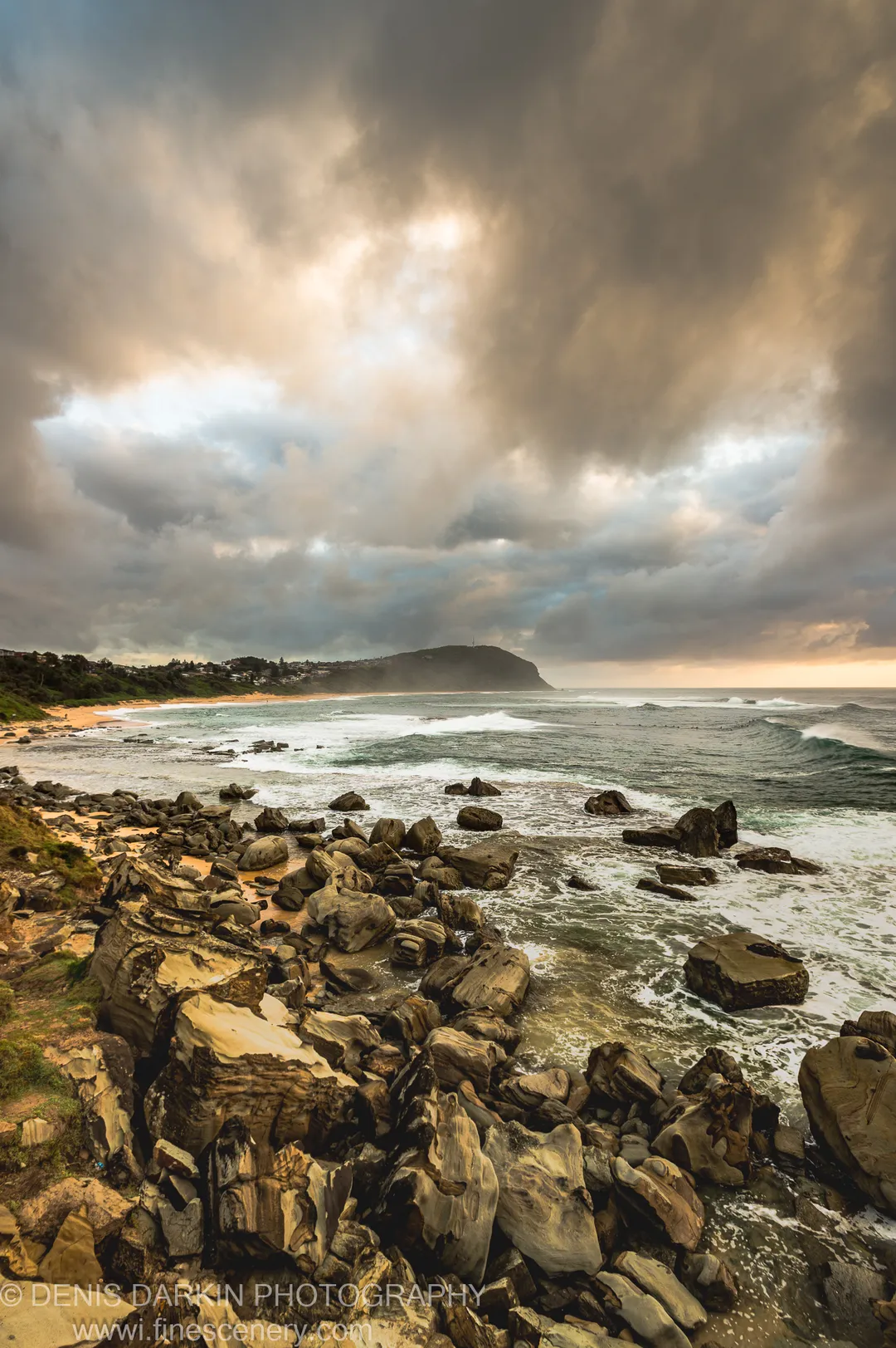 Layered sandstone in the foreground at Spoon Bay, with a crescent beach and headland under heavy cloud on a summer morning.