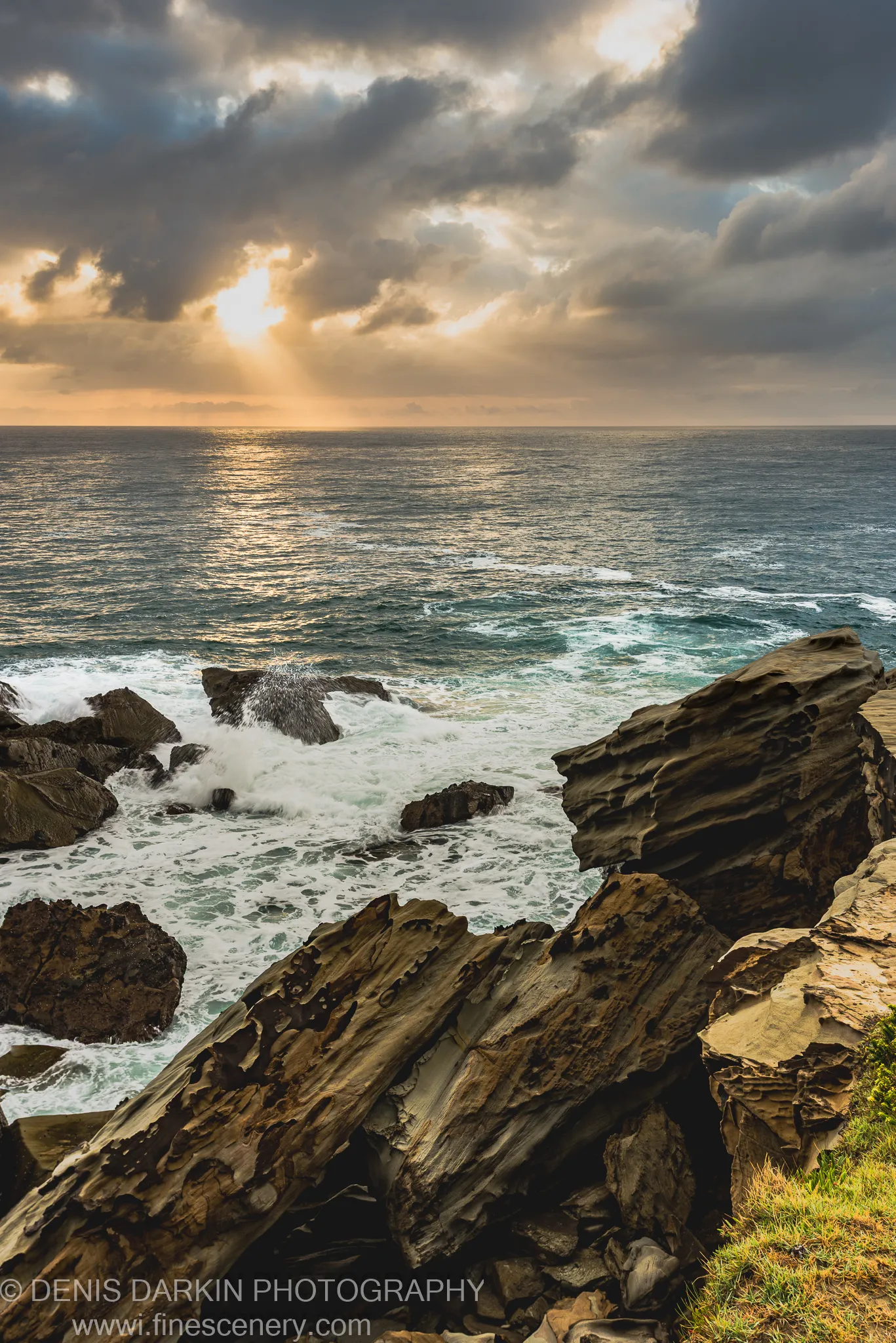 Sunlight breaks through cloud cover above layered sandstone at Spoon Bay, casting light across the ocean on a summer morning.