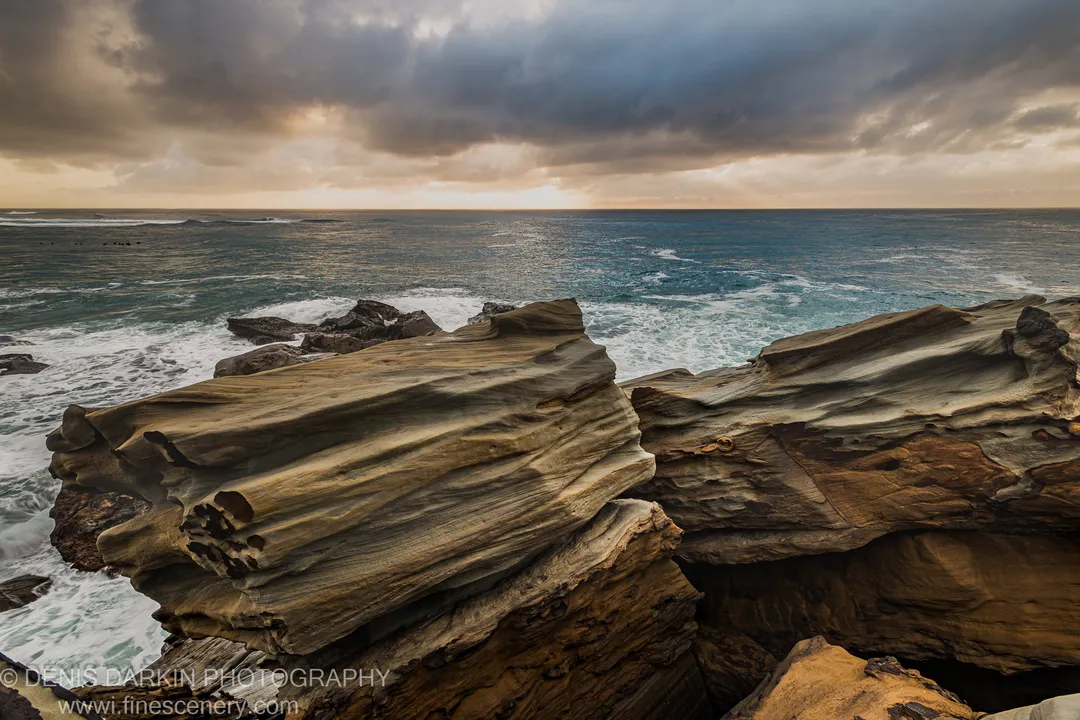 Sculpted ochre sandstone layers at Spoon Bay with wave foam at the rock base and storm clouds above on a summer morning.