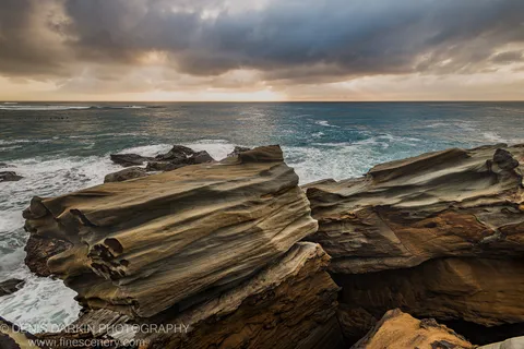 Sculpted ochre sandstone layers at Spoon Bay with wave foam at the rock base and storm clouds above on a summer morning.
