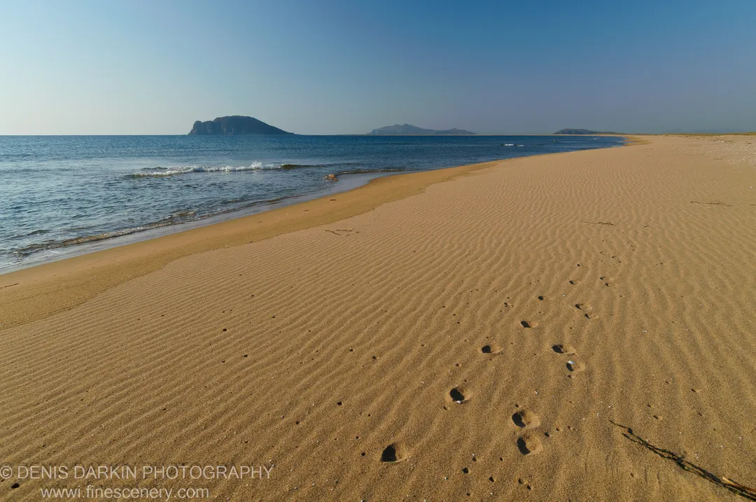 Wind-swept sand ripples cover the beach at Falshivy Poyma, with dark rocky islands visible through haze beyond the gentle surf.