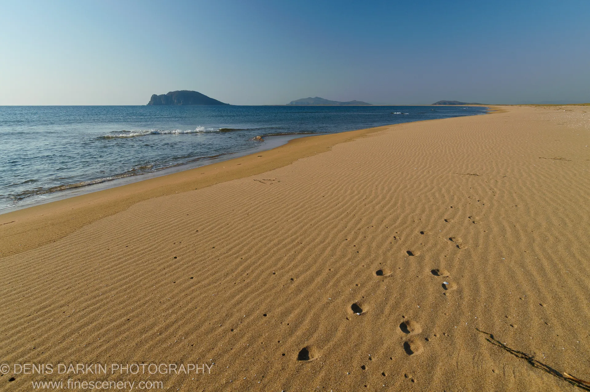 Wind-swept sand ripples cover the beach at Falshivy Poyma, with dark rocky islands visible through haze beyond the gentle surf.