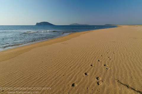 Wind-swept sand ripples cover the beach at Falshivy Poyma, with dark rocky islands visible through haze beyond the gentle surf.