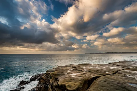 A stratified rock platform at Spoon Bay extends toward a deep blue ocean under backlit clouds on a summer morning.