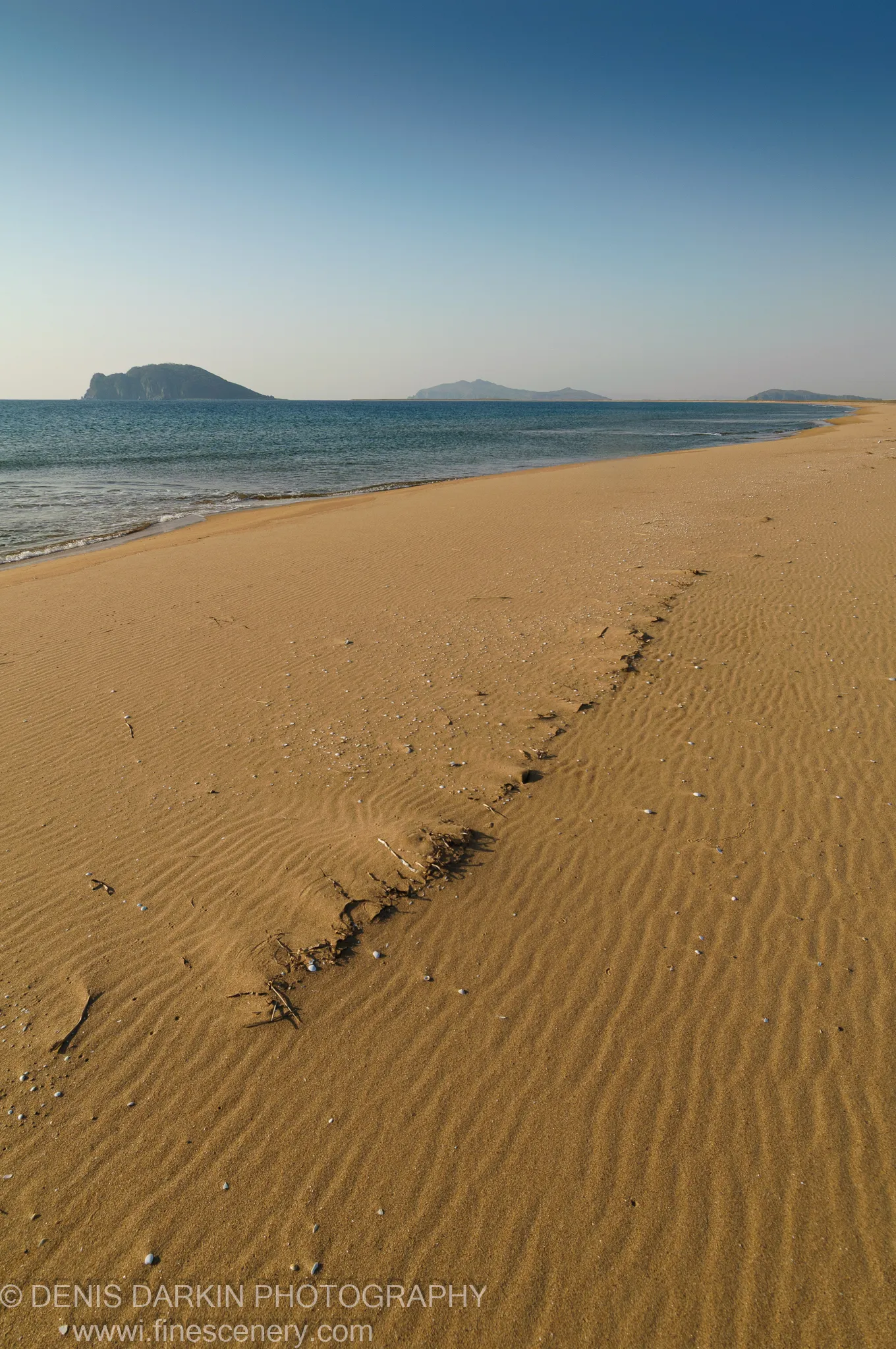 A diagonal line of driftwood and shells crosses wind-rippled sand at Falshivy Poyma, with calm water and distant hazy islands beyond.
