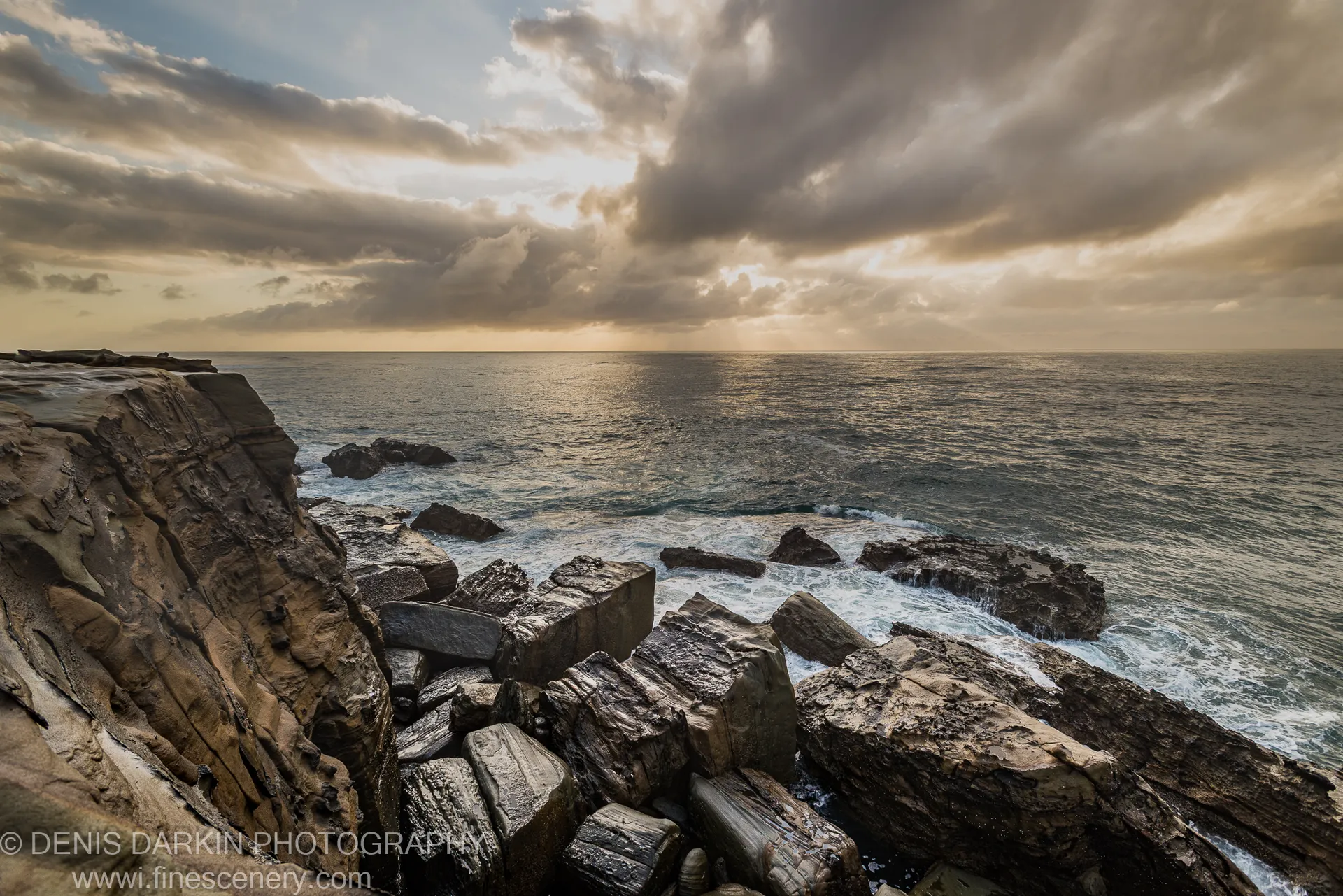 Fractured angular sandstone slabs at Spoon Bay stretch toward the Central Coast horizon, with storm clouds and breaking waves visible in the morning light.