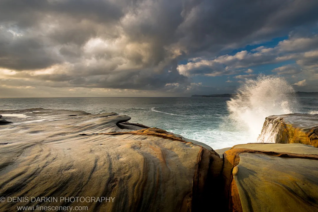 Iron-stained sandstone with swirling surface patterns at Spoon Bay, Central Coast, as a breaking wave sends white spray against the headland cliff face.