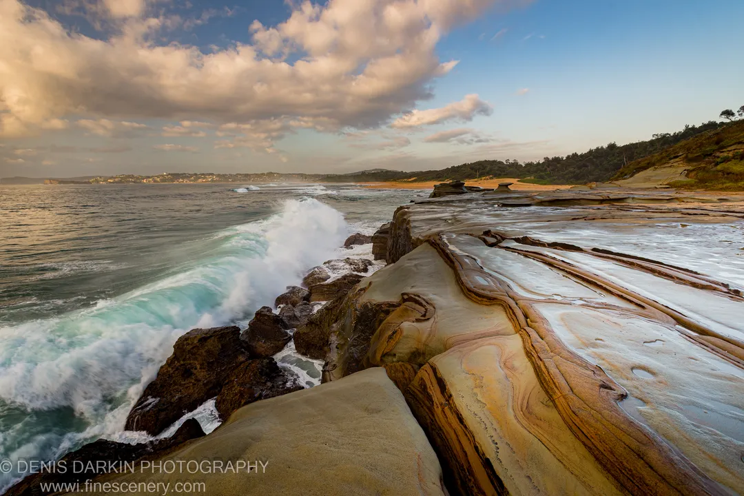 Swirling sandstone layers at Spoon Bay, Central Coast, with sea foam collecting in rock hollows and a breaking wave beyond.