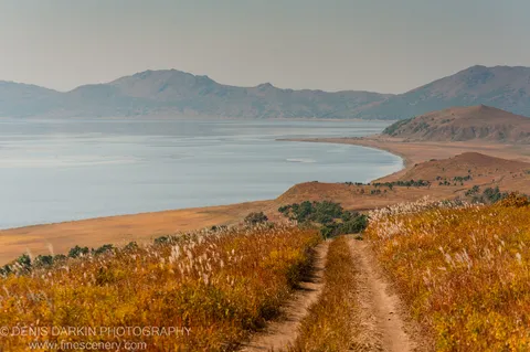 A dirt track winds through tall golden grasses at Falshivy Poyma, leading to calm water and hazy mountains on the horizon.