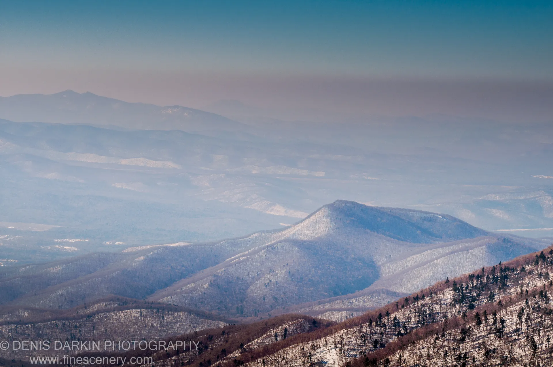Layered mountain ridges at Vorobey Mountain fade into soft blue haze, with a conical dark peak visible and snow-dusted slopes in the foreground, January 2012.