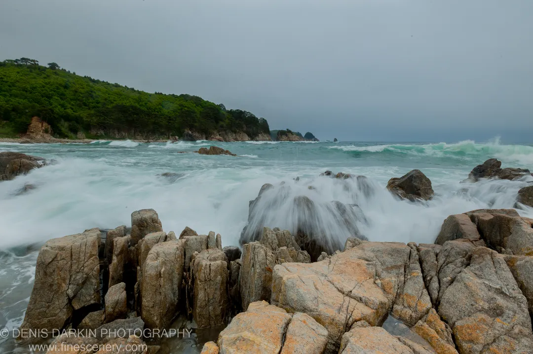 Lichen-covered granite boulders at the Far East Marine Reserve stand against motion-blurred surf while a forested headland recedes into the grey sea.