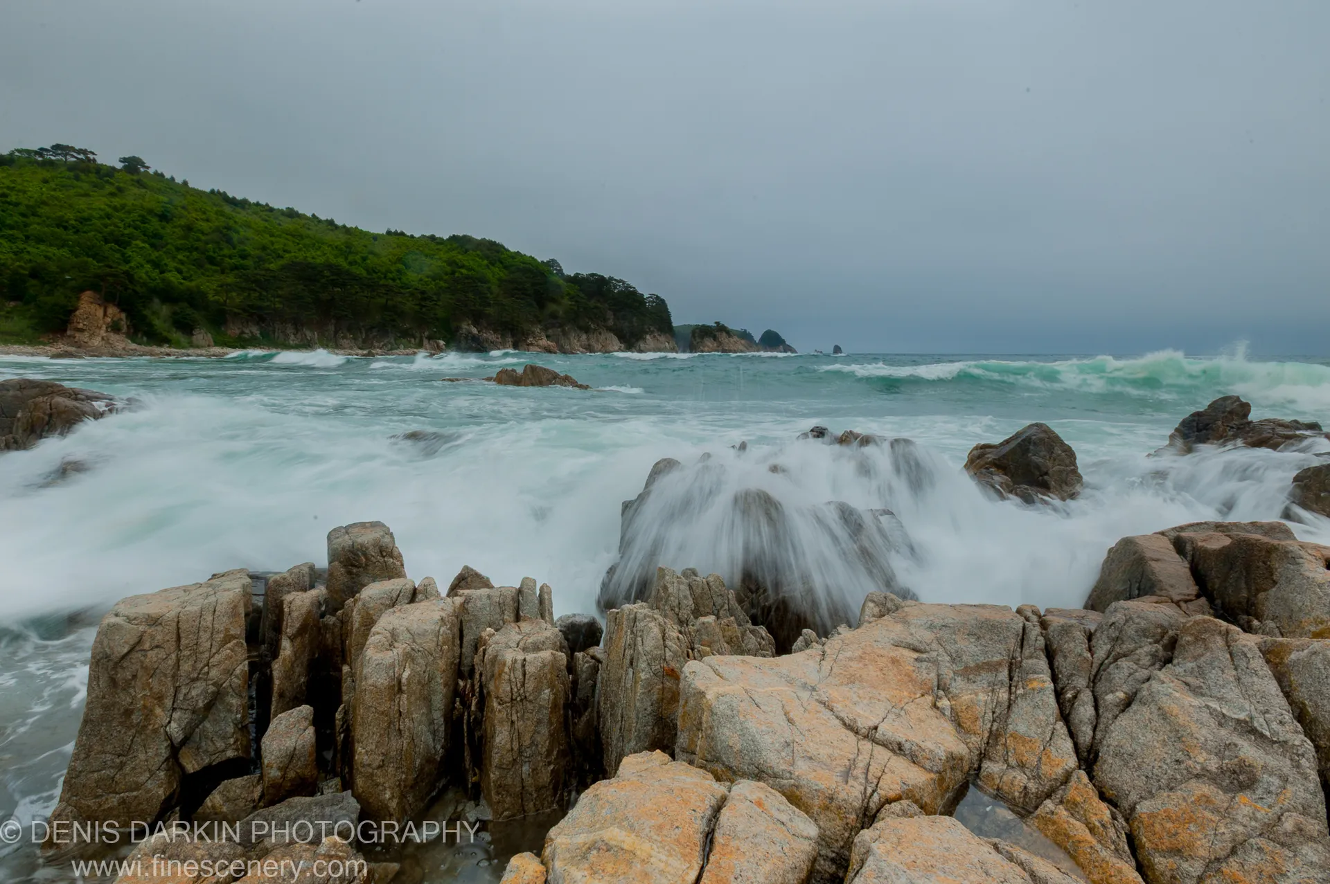 Lichen-covered granite boulders at the Far East Marine Reserve stand against motion-blurred surf while a forested headland recedes into the grey sea.