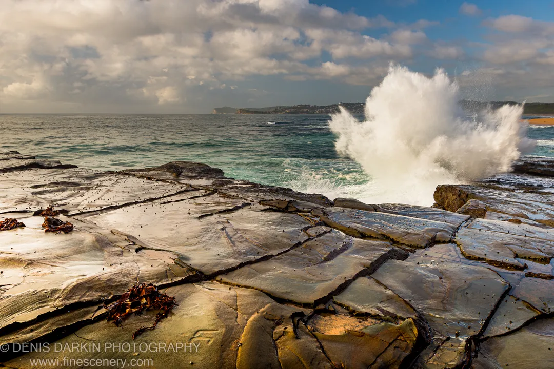A wave crashes against the layered rock at Spoon Bay, Central Coast, sending white spray skyward in summer morning light.