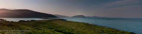 A grassy headland slopes toward a calm bay at sunset near Duhovo, with layered coastal ridges receding into the warm haze.