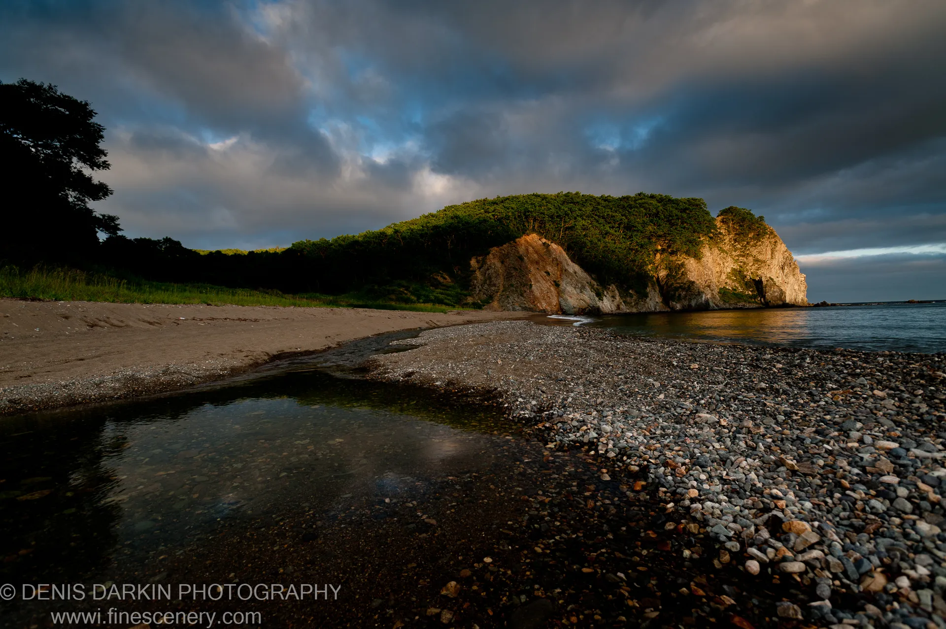Warm evening light catches the forested cliff face at Kamenka as heavy storm clouds build above the pebble shore.