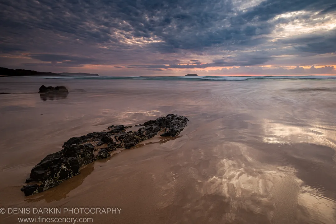 Jagged rock formation in the foreground, with dawn waves and an offshore island along the Gold Coast shoreline.