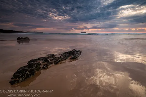 Jagged rock formation in the foreground, with dawn waves and an offshore island along the Gold Coast shoreline.