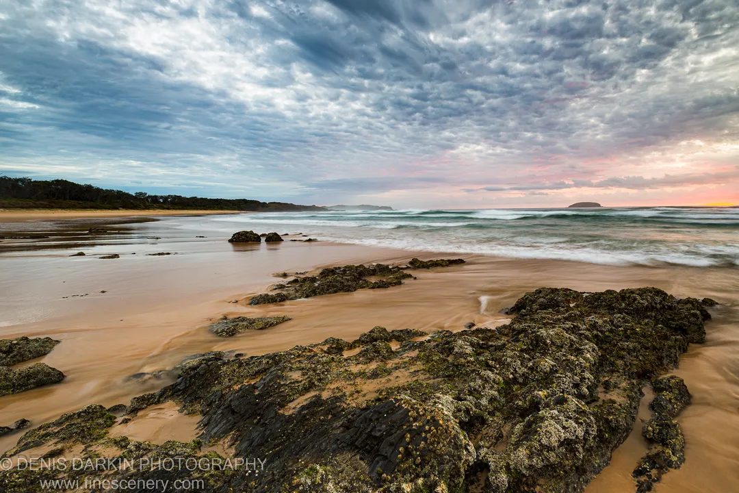 Barnacle-covered rock shelf in the foreground at low tide, with a streaked mackerel sky and a distant headland along the Queensland coast.