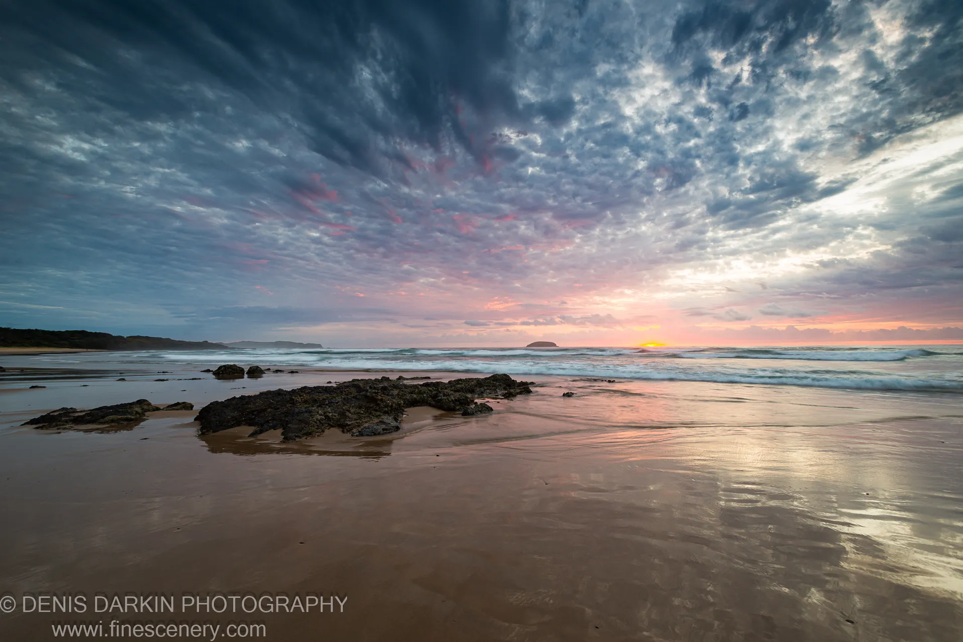Wet sand reflects the pink and blue hues of the dawn sky above dark moss-covered coastal rocks, Gold Coast, Queensland.