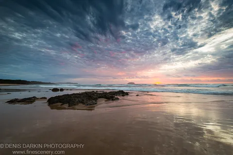 Wet sand reflects the pink and blue hues of the dawn sky above dark moss-covered coastal rocks, Gold Coast, Queensland.