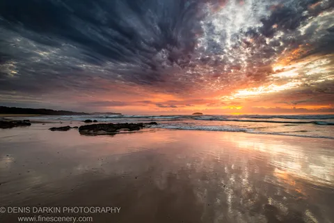 Dark basalt rocks and wet sand reflecting a purple and orange dawn sky, with gentle surf rolling toward the Gold Coast shore.