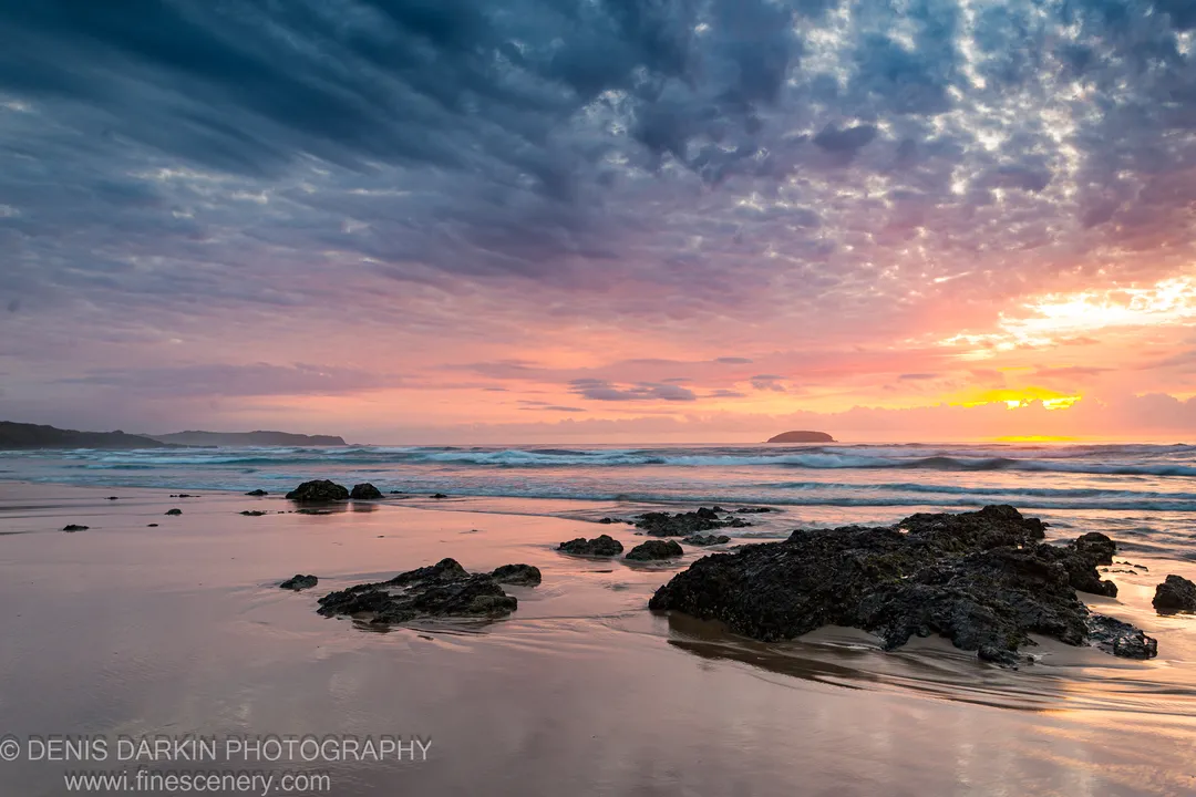 Wet sand reflects pink and orange dawn hues, with dark basalt rocks scattered along the shoreline and streaked clouds overhead at Gold Coast.