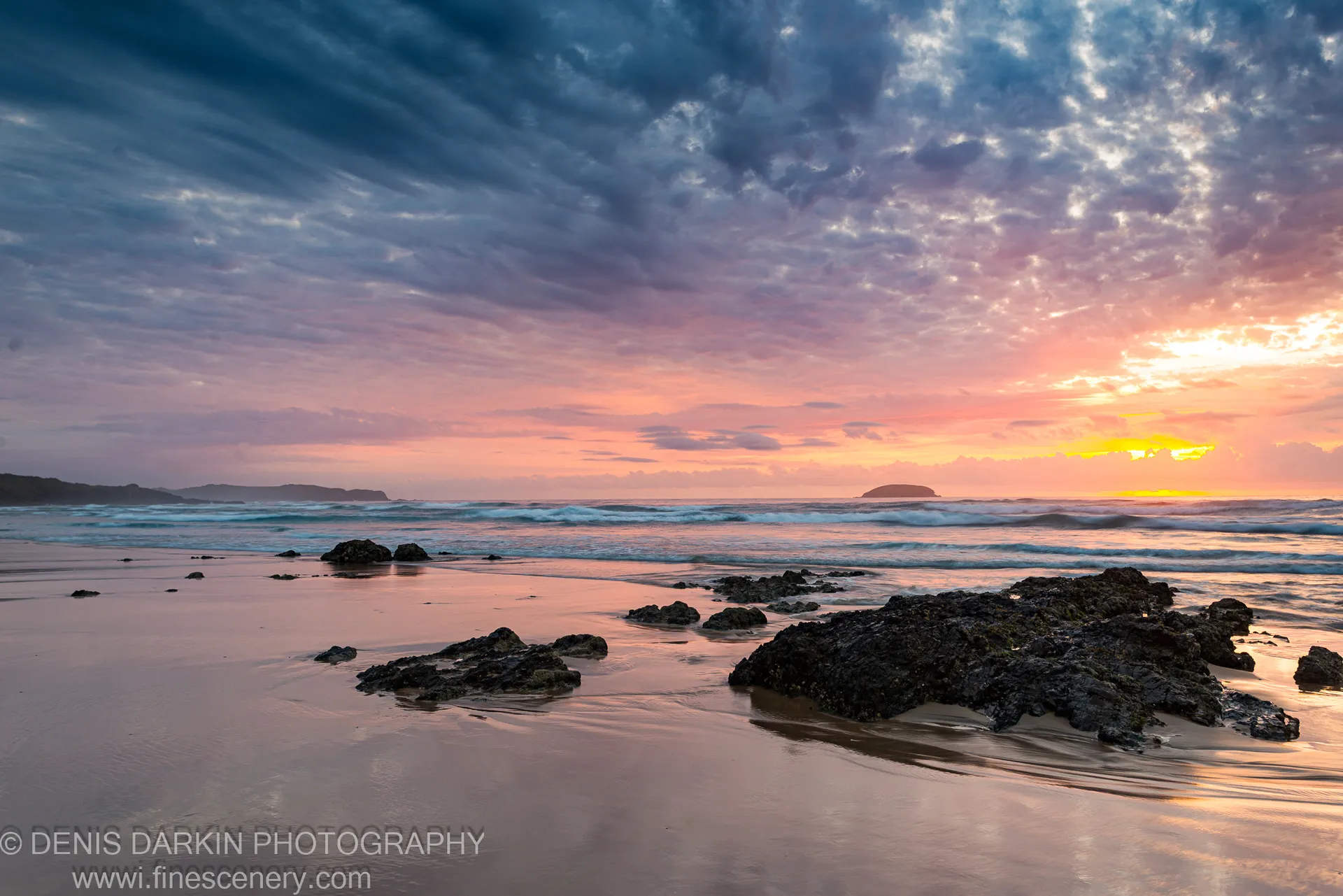Wet sand reflects pink and orange dawn hues, with dark basalt rocks scattered along the shoreline and streaked clouds overhead at Gold Coast.