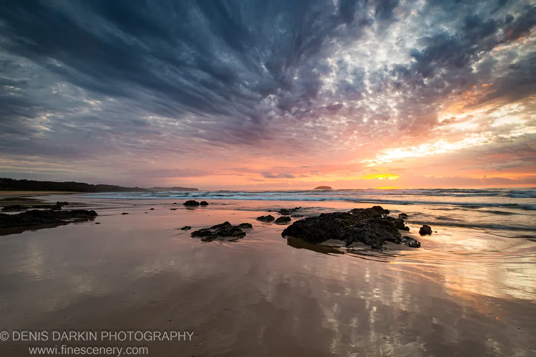 Streaked dawn clouds above a wet mirror beach, dark jagged rocks in the foreground, and a distant headland on the Queensland coast.